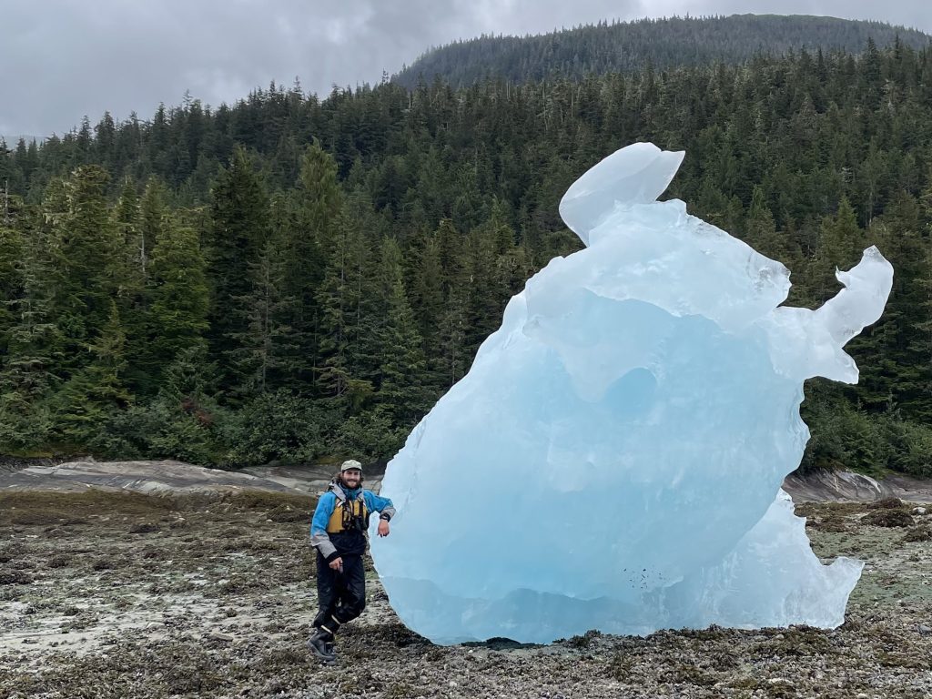 Matt Brodsky in U.S. Forest Service ranger gear standing beneath a towering iceberg in the Tracy Arm–Ford’s Terror Wilderness during patrol.